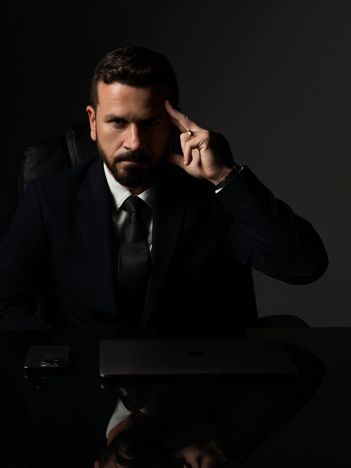 Ed Dalcin sentado à mesa, de terno escuro e gravata, com a mão apoiada na têmpora e olhando para a câmera; à frente, um notebook fechado e um celular
